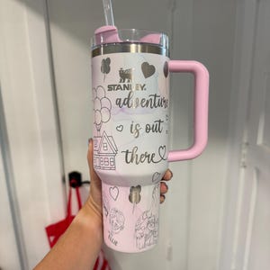 May include: A white and pink Stanley tumbler with a pink handle and lid. The tumbler features the text "adventure is out there" with illustrations of balloons, a house, and characters. A clear straw is inserted into the lid.