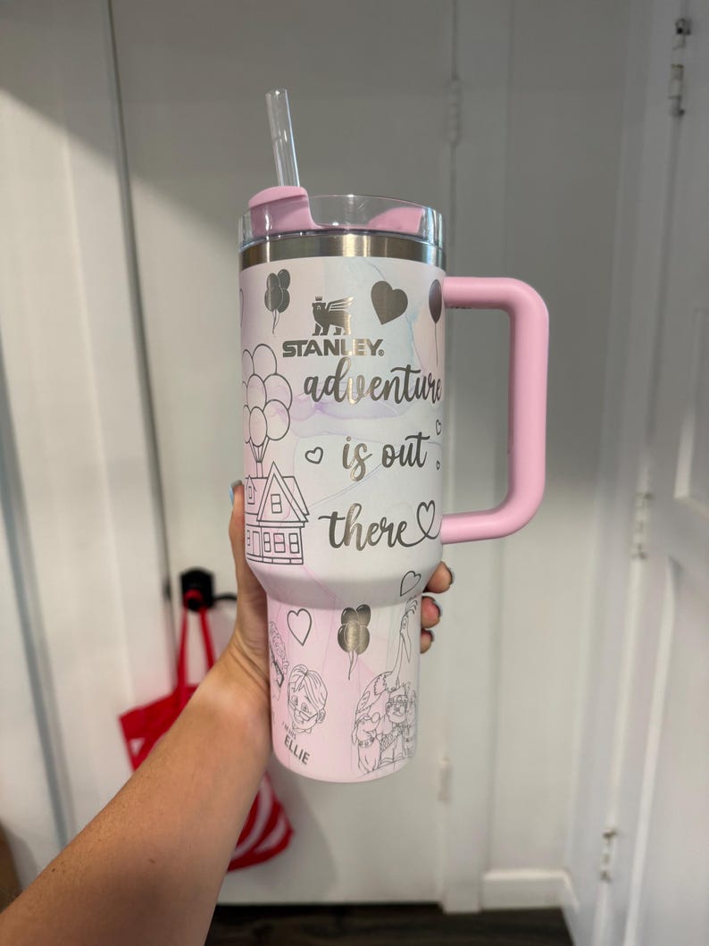 May include: A white and pink Stanley tumbler with a pink handle and lid. The tumbler features the text "adventure is out there" with illustrations of balloons, a house, and characters. A clear straw is inserted into the lid.