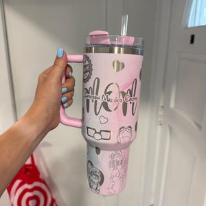 May include: A pink and white tumbler with a handle, lid, and straw. The tumbler features the word "Mom" in silver script, along with names and drawings. The handle and lid are solid pink.