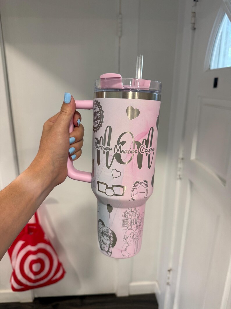May include: A pink and white tumbler with a handle, lid, and straw. The tumbler features the word "Mom" in silver script, along with names and drawings. The handle and lid are solid pink.