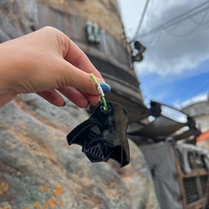 May include: A black plastic keychain shaped like Darth Vader's helmet. The keychain is attached to a green and silver string.