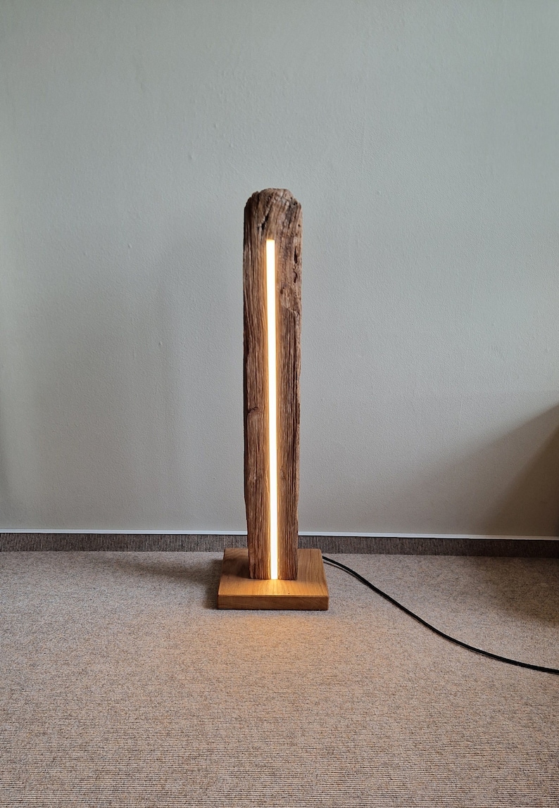 May include: A wooden floor lamp with a natural wood base and a tall, rectangular wood column. The column has a thin, vertical slit that emits a warm white light.
