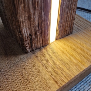 May include: A wooden lamp with a warm, natural wood finish. The lamp has a rectangular base and a tall, cylindrical body. A thin strip of LED light runs vertically down the center of the lamp, illuminating the wood grain.