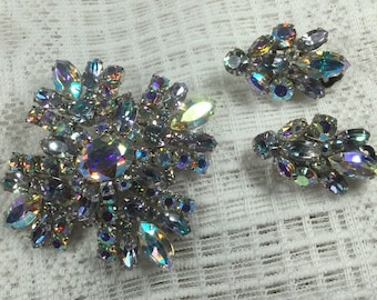 May include: A set of iridescent rhinestone jewelry featuring a large brooch and a pair of earrings. The brooch is a cluster of rhinestones in various shapes and sizes, while the earrings are smaller clusters of rhinestones.