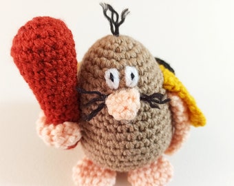 May include: A handmade crochet figure of a cartoon mole. The mole is primarily brown with a peach-colored nose and feet, white eyes, and black whiskers. It holds a red club and has a yellow and black detail on its back.
