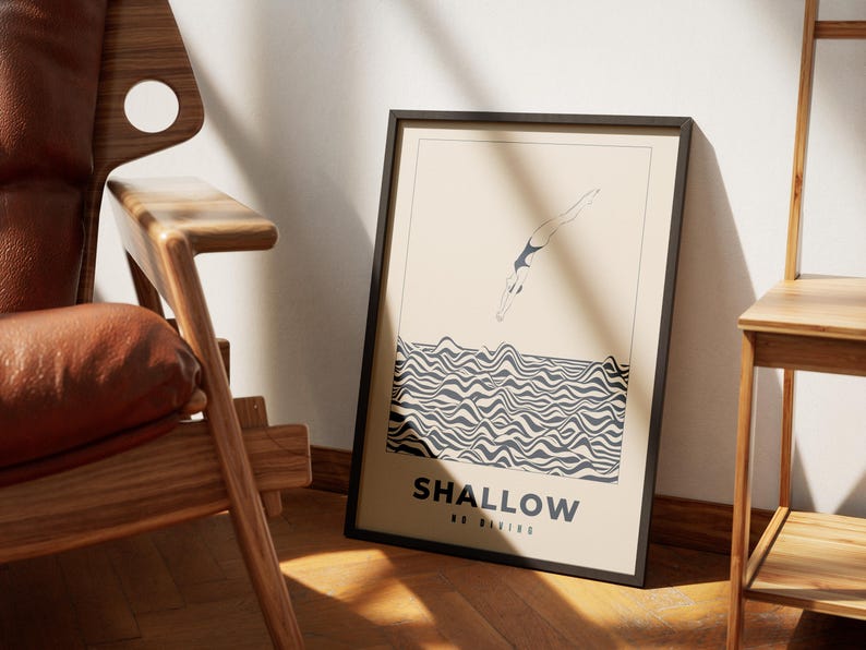 Op de afbeelding: Een zwart ingelijste poster met een beige achtergrond waarop een lijntekening van een persoon te zien is die in golvend zwart-wit water duikt. De tekst "SHALLOW NO DIVING" is in zwart onderaan de poster gedrukt.