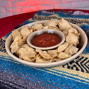 Puede incluir: Un plato de cerámica beige para servir chips y salsa con un tazón más pequeño en el centro para la salsa. El plato está lleno de chips de tortilla y el tazón está lleno de salsa roja. El plato está sobre una manta de rayas de colores.