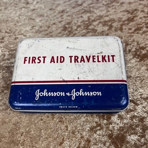 May include: Vintage white metal first aid travel kit with red and blue accents. The kit has the text "FIRST AID TRAVELKIT" in red and "Johnson & Johnson" in blue with the words "PRESS BELOW" in black.