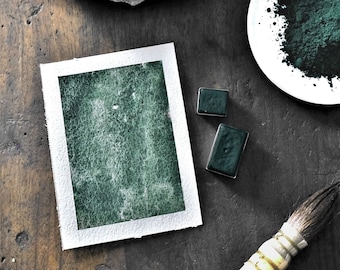 May include: A close-up of a dark green watercolor paint set with a brush, a small rock, and a white dish with a pile of dark green pigment.