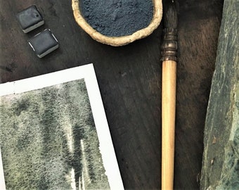 May include: An overhead shot of art supplies on a dark wooden surface. The image includes a small bowl of blue pigment, watercolor paint blocks, a paintbrush, and a watercolor painting. A bowl of amber-colored crystals is also visible.