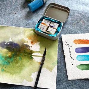Peut inclure: Composition de matériel de peinture à l'aquarelle sur une surface texturée. L'image présente de petits flacons de peinture verte, dorée et bleue, ainsi qu'une boîte métallique contenant des godets d'aquarelle. Un pinceau et des échantillons de couleurs sont également présents.