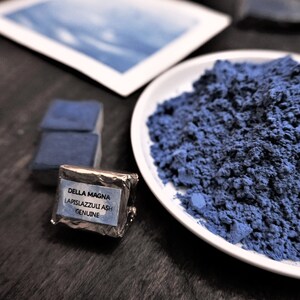 Puede incluir: Un plato blanco lleno de polvo azul se encuentra junto a un peque&ntilde;o paquete etiquetado como "DELLA MAGNA LAPISLAZZULI ASH GENUINE". Tambi&eacute;n se ven dos bloques rectangulares azules y una imagen enmarcada.