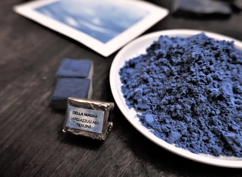 Puede incluir: Un plato blanco lleno de polvo azul se encuentra junto a un peque&ntilde;o paquete etiquetado como "DELLA MAGNA LAPISLAZZULI ASH GENUINE". Tambi&eacute;n se ven dos bloques rectangulares azules y una imagen enmarcada.