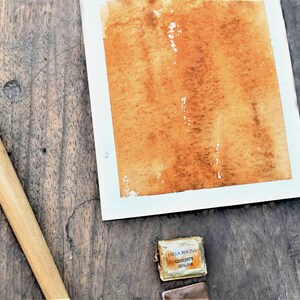 May include: A rectangular piece of white paper featuring a wash of warm, earthy orange-brown watercolour paint. Beside the paper lies a wooden-handled brush with a metal ferrule and a small, foil-wrapped watercolour pan labelled Della Magna Ozocerite.