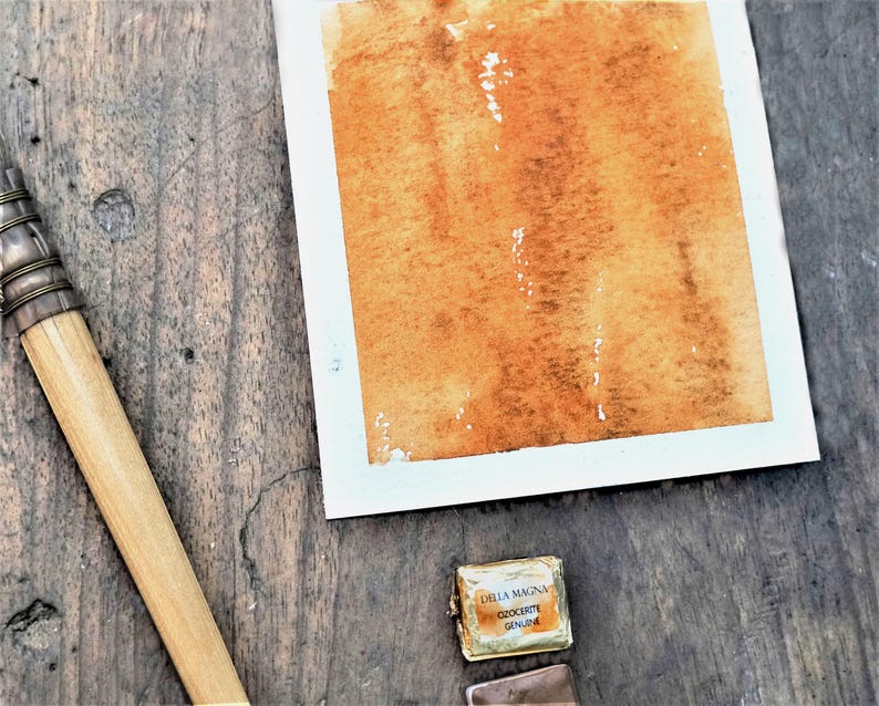 May include: A rectangular piece of white paper featuring a wash of warm, earthy orange-brown watercolour paint. Beside the paper lies a wooden-handled brush with a metal ferrule and a small, foil-wrapped watercolour pan labelled Della Magna Ozocerite.
