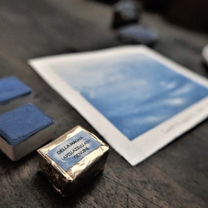 Puede incluir: Una imagen de naturaleza muerta con materiales de acuarela. Dos pastillas de acuarela azules rectangulares y un bloque de pigmento envuelto se colocan cerca de una pintura de acuarela. El pigmento envuelto est&aacute; etiquetado "DELLA MAGNA LAPISLAZZULI ASH GENUINE."