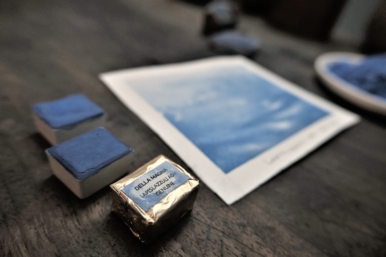 Puede incluir: Una imagen de naturaleza muerta con materiales de acuarela. Dos pastillas de acuarela azules rectangulares y un bloque de pigmento envuelto se colocan cerca de una pintura de acuarela. El pigmento envuelto est&aacute; etiquetado "DELLA MAGNA LAPISLAZZULI ASH GENUINE."