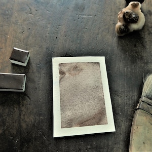 May include: A watercolor painting supplies still life. Two rectangular watercolor paint pans and a watercolor brush are arranged around a small watercolor painting on a wooden surface. The painting has a brown and beige color scheme.