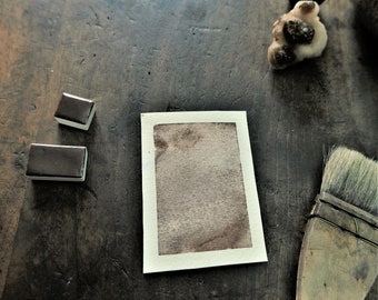 May include: A watercolor painting supplies still life. Two rectangular watercolor paint pans and a watercolor brush are arranged around a small watercolor painting on a wooden surface. The painting has a brown and beige color scheme.