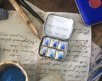 May include: An open tin of Della Magna watercolors, "Historical Blues Set," with six small, wrapped paint squares. A blue tin with a wax seal is nearby. The scene includes a calligraphy pen, a small bowl of blue pigment, and aged paper.