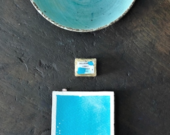 Può includere: Una ciotola in ceramica turchese, un piccolo quadrato di acquerello con la scritta "DELLA MA COBALT BOISE 500" e un foglio di carta per acquerello rettangolare con un lavaggio blu, sono disposti su una superficie scura.