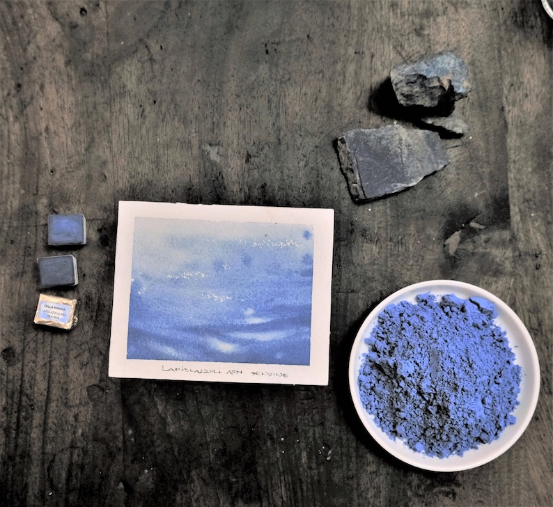 Puede incluir: Una composici&oacute;n de naturaleza muerta con pinturas y pigmentos de acuarela. La imagen incluye una pintura de acuarela con tonos azules, dos peque&ntilde;os bloques de pintura, un plato de pigmento azul y trozos de piedra en bruto. El texto "LAPISLAZULI APH GENUINE" es visible.