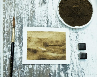 May include: A close-up of a watercolor painting of a brown landscape. The painting is on a white sheet of paper. There is a paintbrush, a small dish of brown paint powder, and two small squares of brown paint on a white wooden surface.