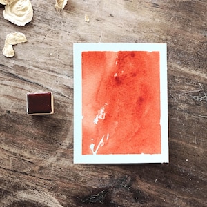 May include: A rectangular piece of white paper featuring a vibrant orange watercolour wash with visible brush strokes and texture, positioned next to a small, dark red rectangular watercolour paint pan on a weathered wooden surface.