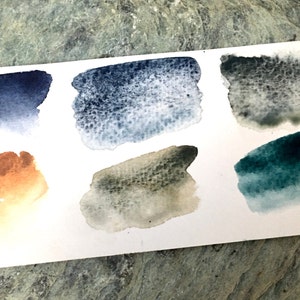 K&ouml;nnte beinhalten: Eine Aquarell-Farbmusterkarte mit sechs verschiedenen Blaut&ouml;nen, Gr&uuml;nt&ouml;nen und Braunt&ouml;nen. Die Farbmuster sind rechteckig geformt und in zwei Reihen mit jeweils drei Mustern angeordnet.