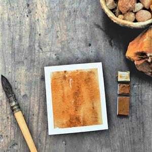 May include: A rectangular piece of paper featuring a textured watercolour wash in shades of burnt orange and brown. Beside the paper are three small rectangular watercolour paint pans in varying brown tones and a wooden handled paintbrush.