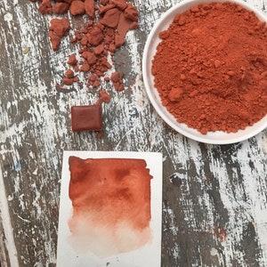 Könnte beinhalten: Draufsicht auf Kunstmaterialien. Zerbröseltes rotbraunes Pigment liegt neben einer weißen Schale mit demselben Pigment. Ein kleiner Pigmentblock und eine Aquarellprobe sind ebenfalls vorhanden.