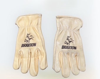 Puede incluir: Un par de guantes de trabajo de cuero de color beige claro. Cada guante presenta un gráfico negro de un vaquero en un toro sobre el nombre "BRAXTON". Los guantes están diseñados para el trabajo y la protección.