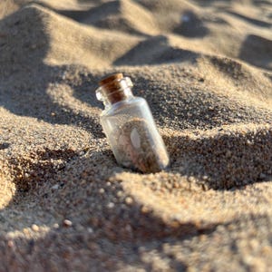 Peut inclure: Une petite bouteille en verre transparent avec un bouchon en liège, remplie de sable et de petits coquillages. La bouteille est posée sur du sable, avec des ombres créées par le soleil. L'ensemble évoque un thème de plage.