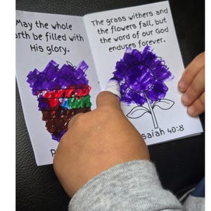 May include: A children's book with drawings and text. One page has a drawing of a purple flower and the text "The grass withers and the flowers fall, but the word of our God endures forever." The other page has a drawing of a purple object and the text "May the whole earth be filled with His glory."