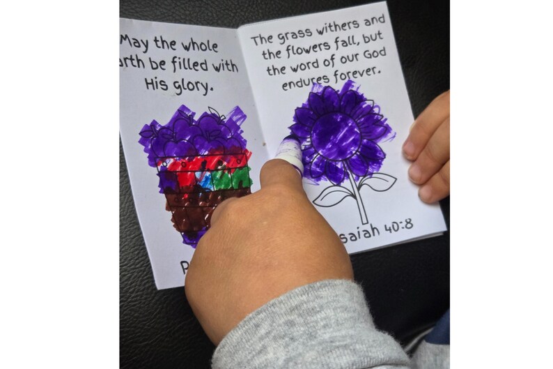 May include: A children's book with drawings and text. One page has a drawing of a purple flower and the text "The grass withers and the flowers fall, but the word of our God endures forever." The other page has a drawing of a purple object and the text "May the whole earth be filled with His glory."