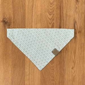 Puede incluir: Un pañuelo para perro azul claro con un patrón de triángulos blancos. El pañuelo tiene una etiqueta de cuero marrón con el texto "ARLO & CO."