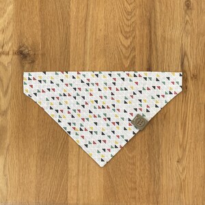 Puede incluir: Un pañuelo blanco con un patrón geométrico de triángulos rojos, amarillos, verdes y negros. El pañuelo tiene una etiqueta marrón con el texto "ARLO & CO" en ella.