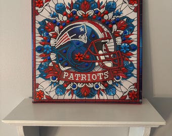 May include: Square canvas art featuring a football helmet design. The helmet is blue and white, with the word "PATRIOTS" in white. The artwork is framed with red and blue accents, and floral patterns.