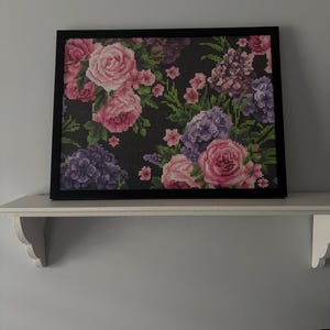 Peut inclure: Tableau floral encadré avec des roses roses et violettes, des hortensias et d'autres fleurs sur un fond sombre. L'œuvre est dans un cadre noir et repose sur une étagère blanche.