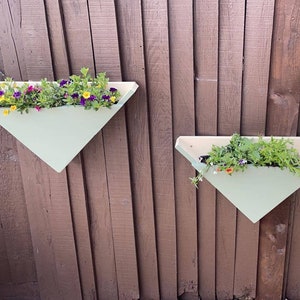 May include: Two white triangular wooden planters with green plants and purple, yellow, and orange flowers hanging on a brown wooden fence.