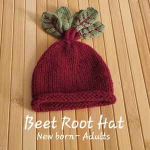 May include: A red knitted beet root hat with green knitted leaves on top. The hat is on a wooden surface. "Beet Root Hat New born + Adults" is written below the hat.