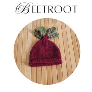 May include: A knit hat pattern titled BEETROOT by KNITWINDOW for an intermediate knitter. The image features a deep burgundy knitted hat with a rolled brim and green knitted leaves attached to the top, resembling a beetroot vegetable.