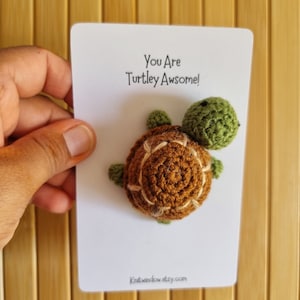 May include: A white card with the text "You Are Turtley Awesome!" and a small crocheted turtle in brown and green colours. The card has the website "Knitwindowetsy.com" printed on the bottom.