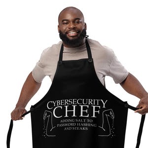 May include: Black apron with the text "CYBERSECURITY CHEF" in white, along with the phrase "ADDING SALT TO PASSWORD HASHING AND STEAKS." The apron has black ties and is worn by a person.