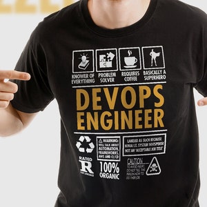 May include: Black t-shirt with a gold graphic design that says "DevOps Engineer". The graphic includes icons for a computer, a wrench, a coffee cup, and a superhero. The text "Rated R 100% Organic" is also included in the graphic.
