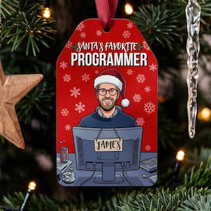 May include: A rectangular red Christmas ornament featuring a cartoon illustration of a person wearing a Santa hat and glasses, sitting at a computer. The text reads SANTA'S FAVORITE PROGRAMMER and JAMES on the monitor. It hangs from a red ribbon.