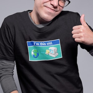 Peut inclure: T-shirt noir avec un motif représentant un globe, un ordinateur et le texte "I'm this old." Le motif est dans un rectangle bleu et vert. La personne porte des lunettes et fait un pouce levé.