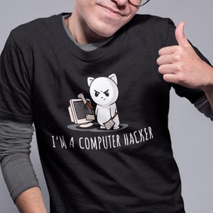 Peut inclure: T-shirt noir avec un dessin d'un chat blanc en colère tenant un marteau et se tenant à côté d'un ordinateur cassé. Le texte "I'M A COMPUTER HACKER" est en dessous. La personne porte des lunettes.