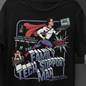 Può includere: T-shirt nera con un'illustrazione in stile fumetto. Un uomo in posa da supereroe tiene un computer portatile, con la scritta "The Family Tech Support Man". Altri testi includono "Ctrl+Alt+Defeat" e "Switch it off... switch it back on."