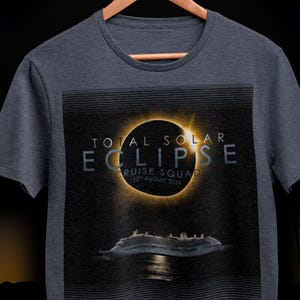 May include: Navy heather t-shirt with a graphic of a total solar eclipse and a cruise ship. Text reads "TOTAL SOLAR ECLIPSE CRUISE SQUAD 12th AUGUST 2024". The t-shirt is displayed on a wooden hanger.
